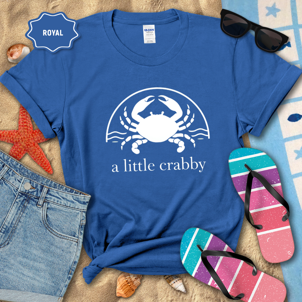 Blue t-shirt with a crab graphic and text, surrounded by beach accessories on sand.