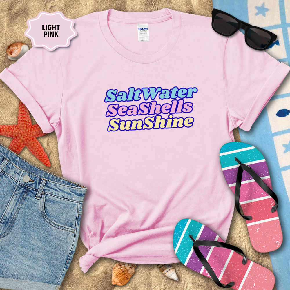 Light pink t-shirt with 'SaltWater SeaShells Sunshine' text, sunglasses, flip-flops, and beach items on sand.