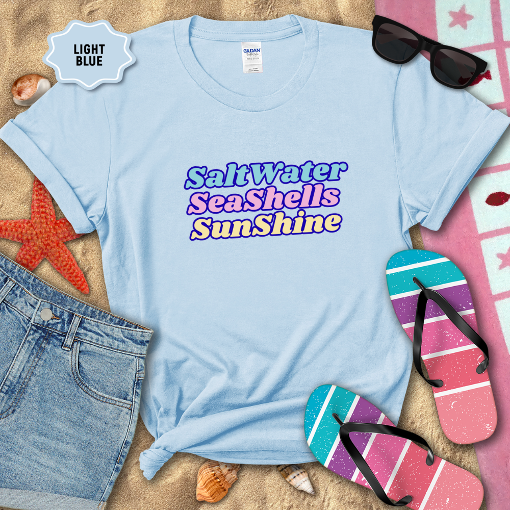 Light blue t-shirt with 'SaltWater SeaShells Sunshine' text, surrounded by beach-themed items on sand.