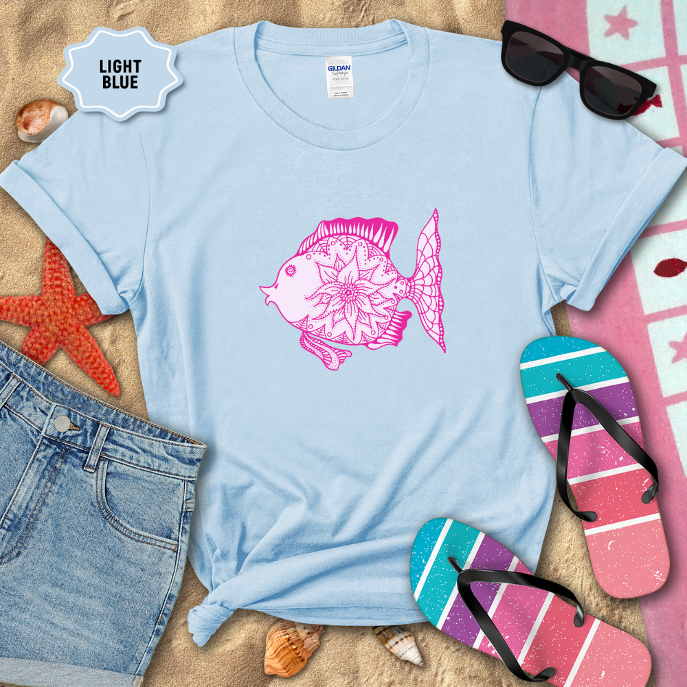 Light blue t-shirt with pink fish design on a sandy background with beach accessories.