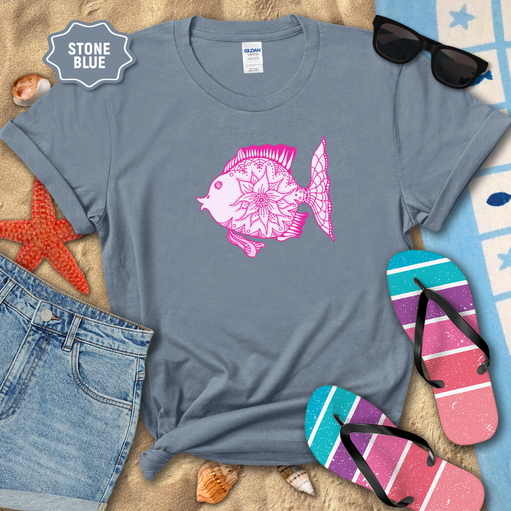 Stone blue t-shirt with pink fish design on a sandy background with beach accessories.