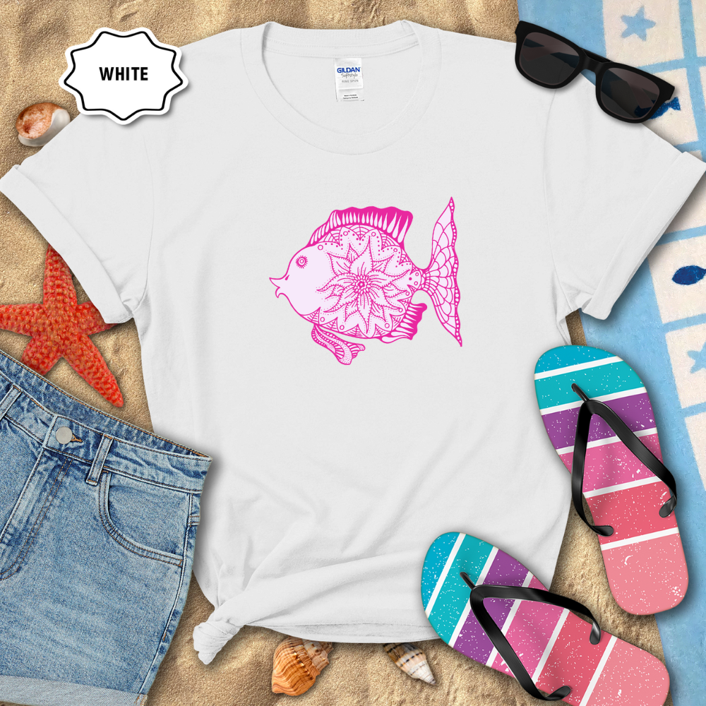 White t-shirt with pink fish design on a sandy surface with beach accessories.