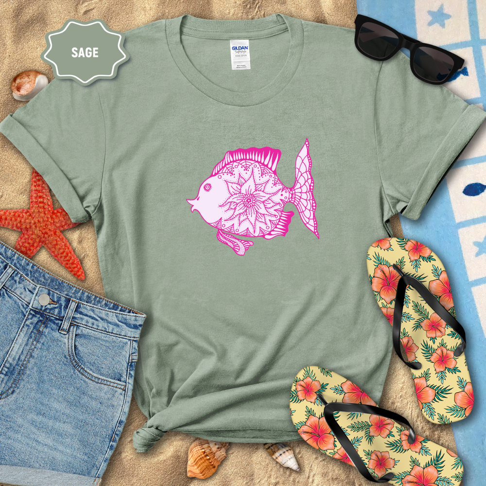 Sage green t-shirt with pink fish design on a sandy surface with beach accessories.