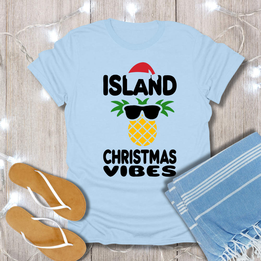 Light blue t-shirt with 'Island Christmas Vibes' design on a wooden surface with flip-flops and a towel.