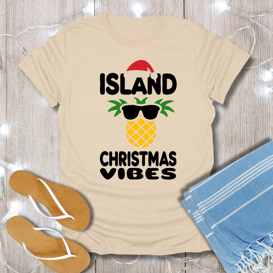 Beige t-shirt with 'Island Christmas Vibes' design on a wooden surface with flip-flops and a towel.