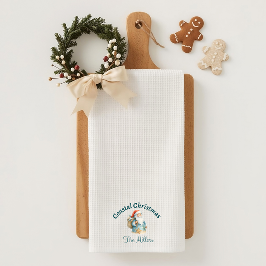 White towel with 'Coastal Christmas' design on a wooden cutting board with a small Christmas wreath and gingerbread cookies.