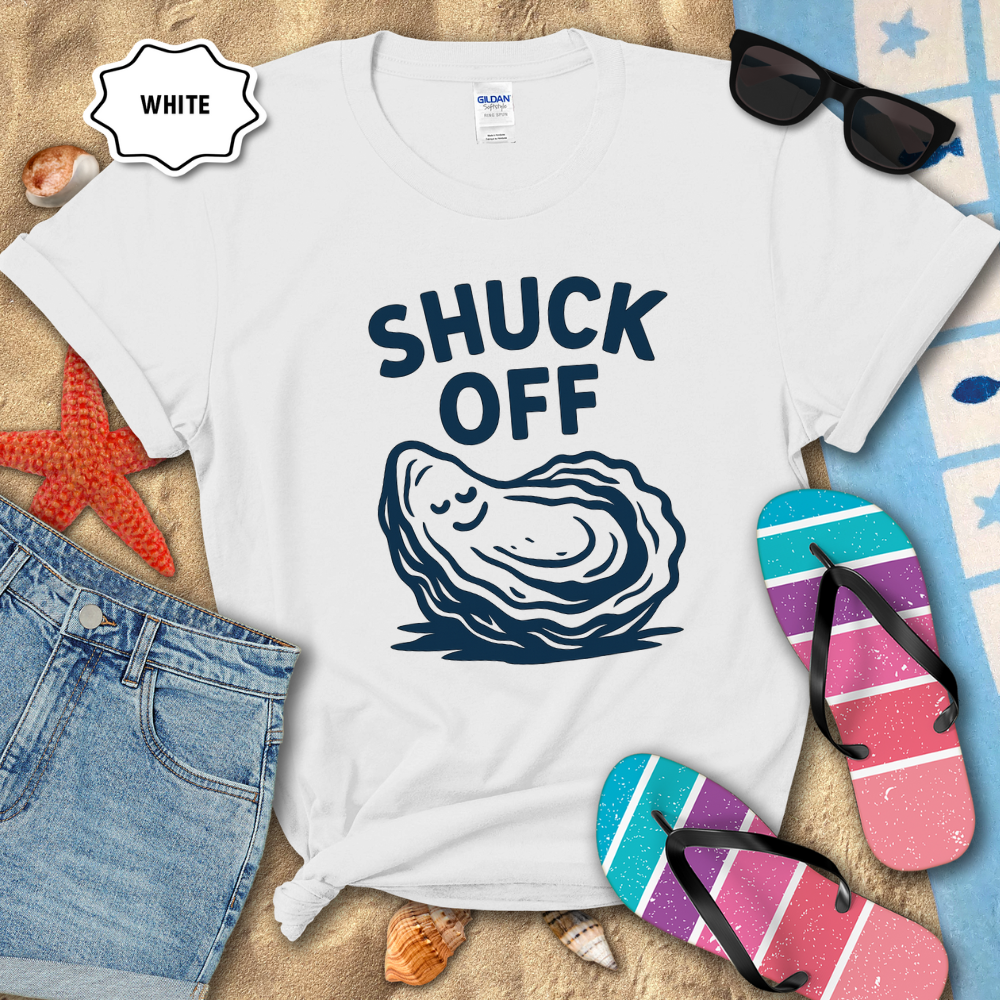 White t-shirt with 'SHUCK OFF' graphic on a sandy surface with beach accessories.