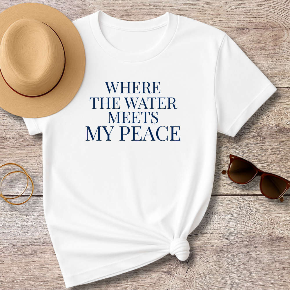 White t-shirt with 'WHERE THE WATER MEETS MY PEACE' text, placed on a wooden surface with a hat and sunglasses.