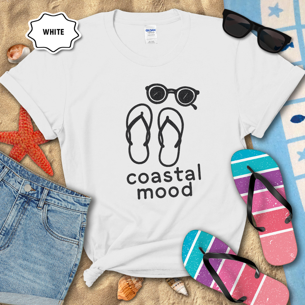 White t-shirt with 'coastal mood' design featuring flip-flops and sunglasses, surrounded by beach accessories.