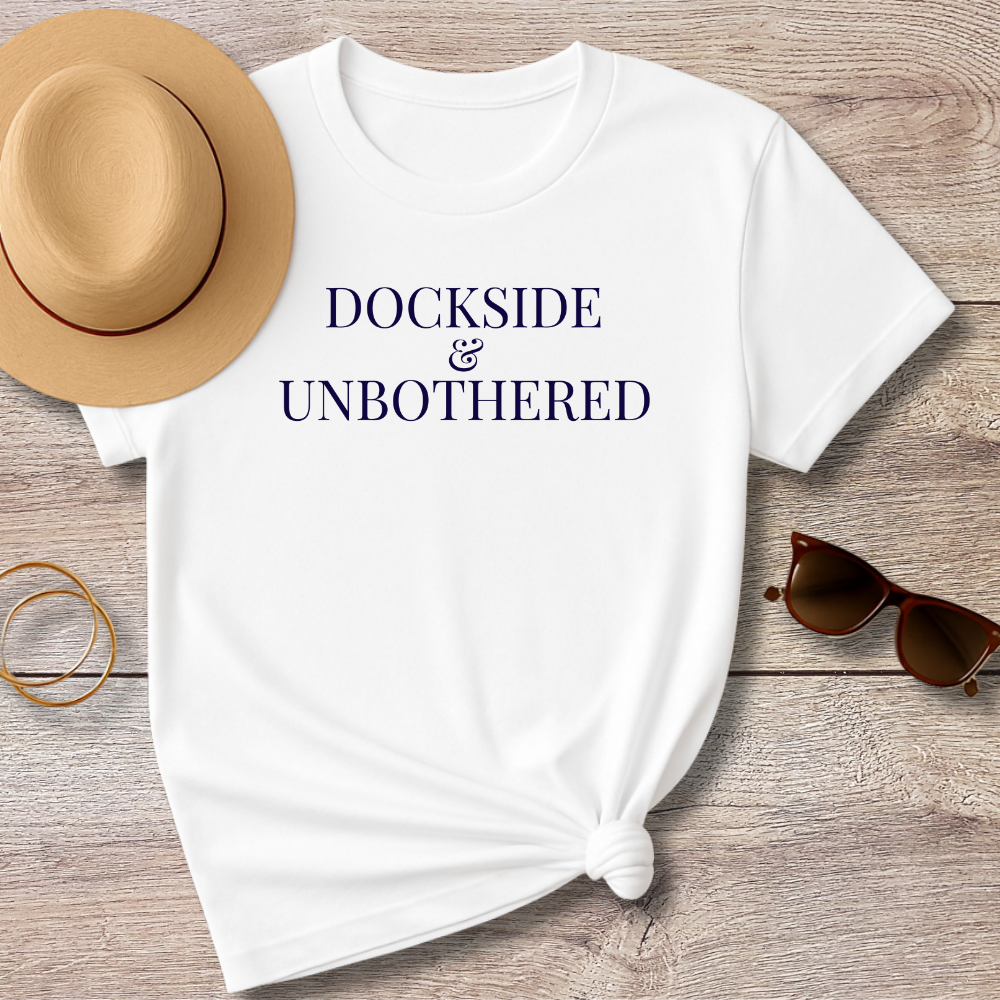 White t-shirt with 'DOCKSIDE & UNBOtherED' text, sun hat, sunglasses, and bracelet on a wooden surface