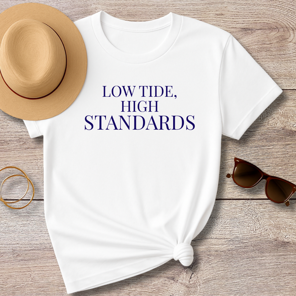 White t-shirt with 'LOW TIDE, HIGH STANDARDS' text on a wooden surface with a hat and sunglasses.
