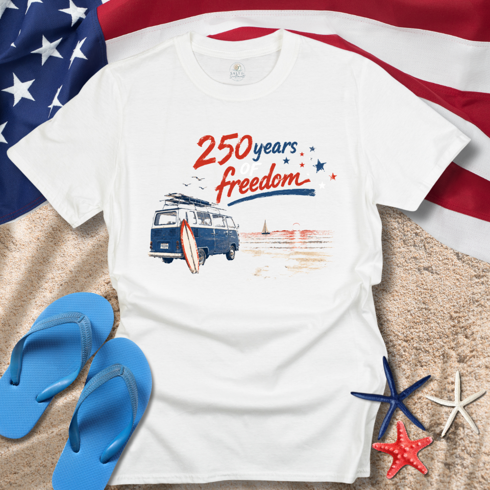 White t-shirt with '250 years freedom' design on a beach setting with American flag, flip-flops, and starfish.
