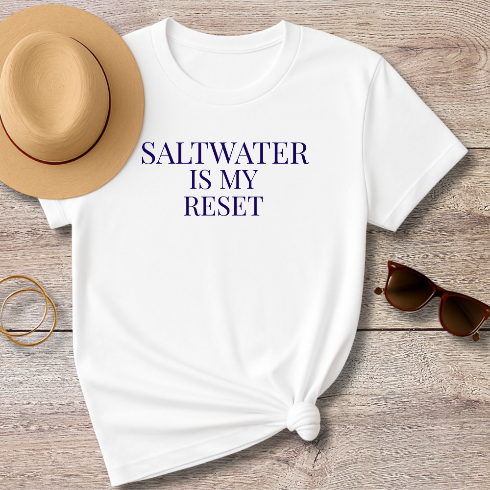 White t-shirt with 'SALTWATER IS MY RESET' text, sun hat, sunglasses, and bracelet on a wooden surface