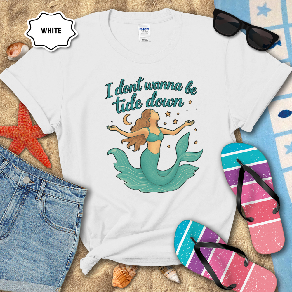 White t-shirt with mermaid graphic and text on a sandy background with beach accessories.