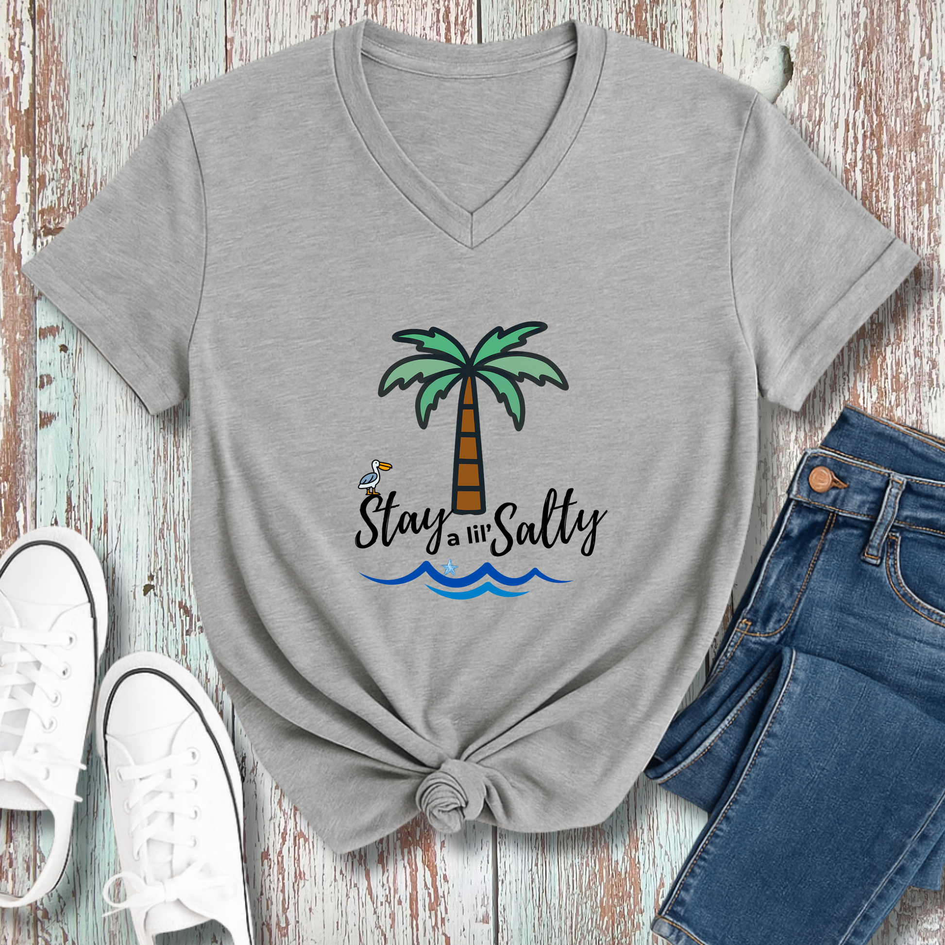 Gray t-shirt with palm tree and 'Stay Salty' design, paired with jeans and sneakers on a wooden surface.