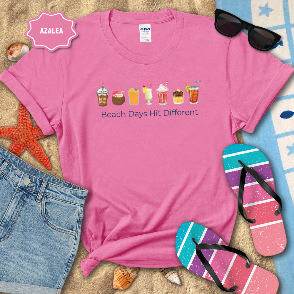 Pink t-shirt with beach-themed graphics and text, surrounded by beach accessories on sand.