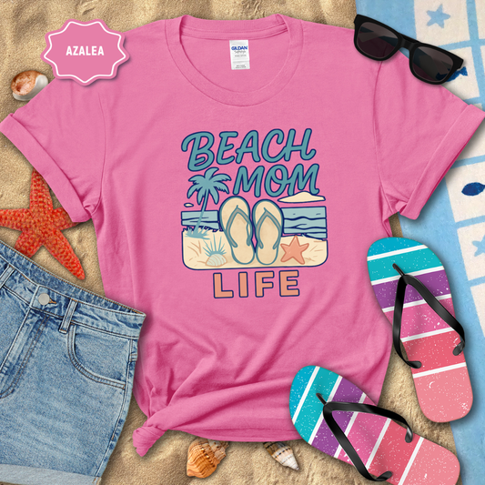 Pink t-shirt with 'Beach Mom Life' design on a beach-themed background with sunglasses, flip-flops, and denim shorts.