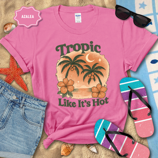 Pink t-shirt with tropical design and text, surrounded by beach accessories on sand.