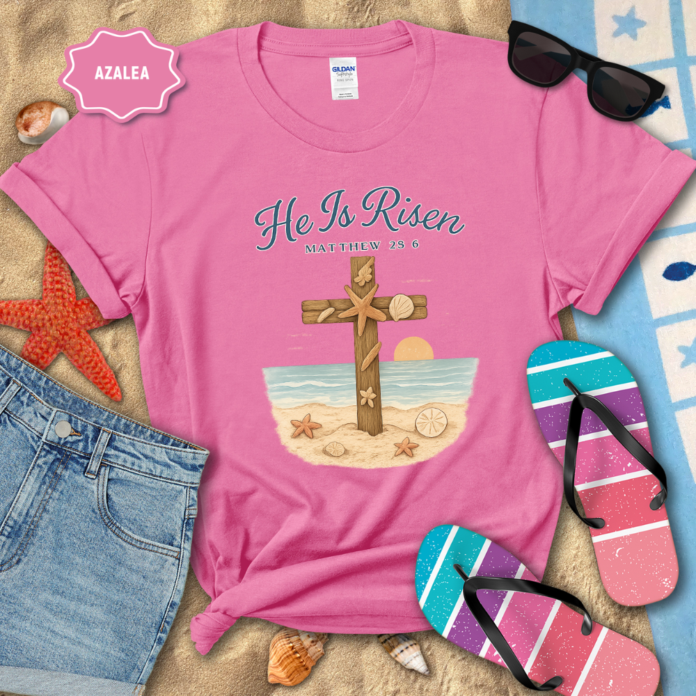 Pink t-shirt with 'He Is Risen' design on a sandy background with beach accessories.