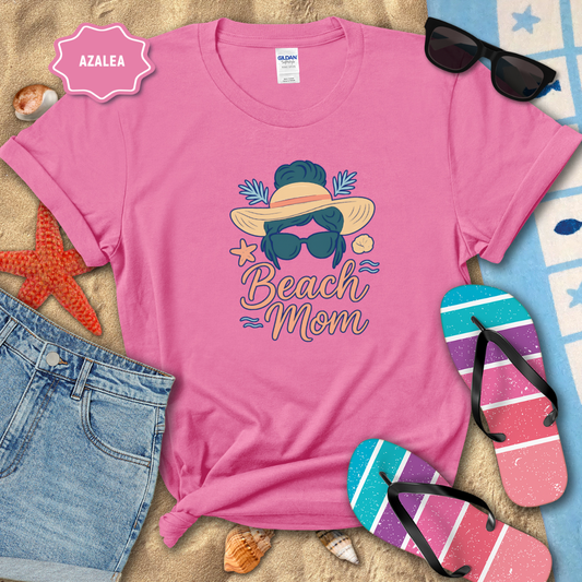 Pink t-shirt with 'Beach Mom' design on a sandy background with sunglasses, flip-flops, and denim shorts.