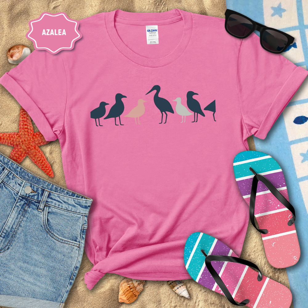 Pink t-shirt with bird design on a beach setting with sunglasses, flip-flops, and denim shorts.