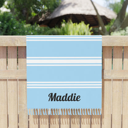 A blue boho beach cloth with white stripes and the name 'Maddie' printed on it, hanging over a wooden fence.