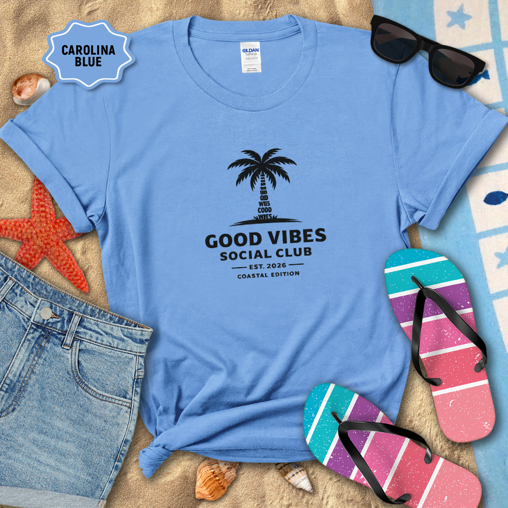 Blue t-shirt with 'Good Vibes Social Club' design on a sandy surface with beach accessories.