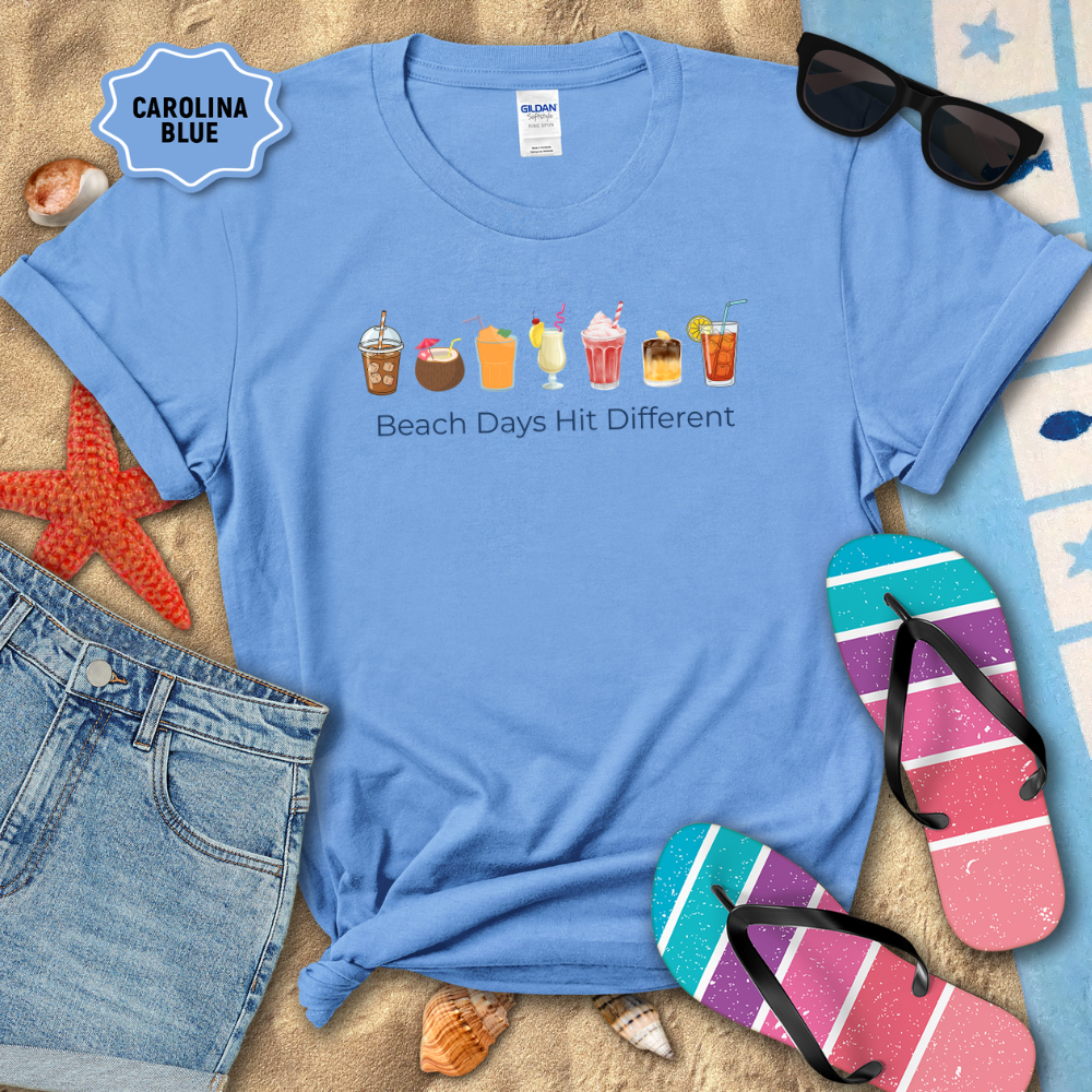Blue t-shirt with beach-themed graphics and text, surrounded by beach accessories on sand.