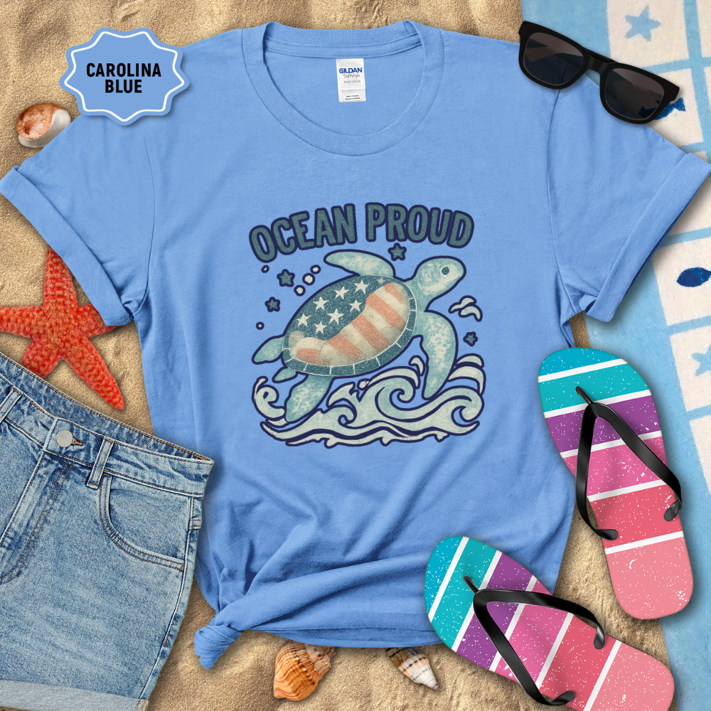 Blue t-shirt with turtle graphic and 'Ocean Proud' text on a beach setting with sunglasses, flip-flops, and starfish.