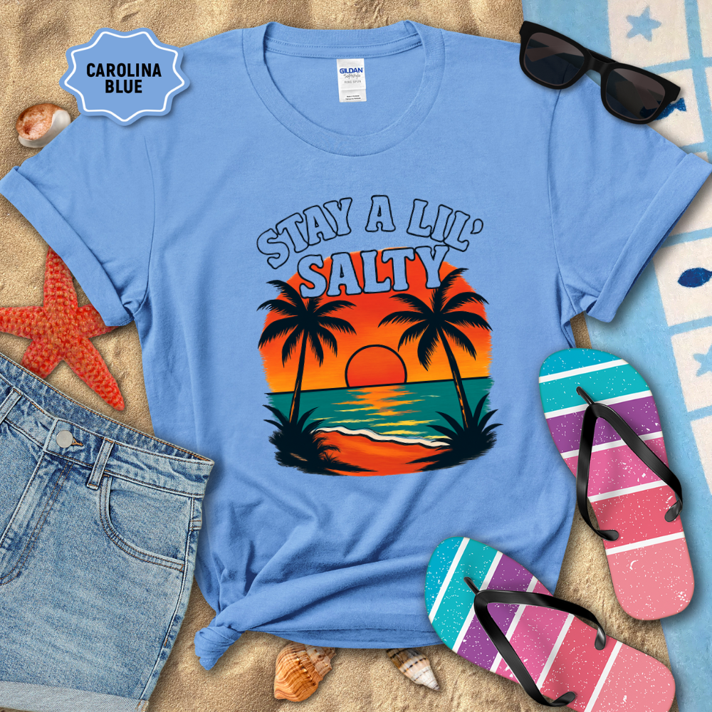 Blue t-shirt with 'Stay a Lil Salty' design, sunglasses, flip-flops, and beach accessories on sand.