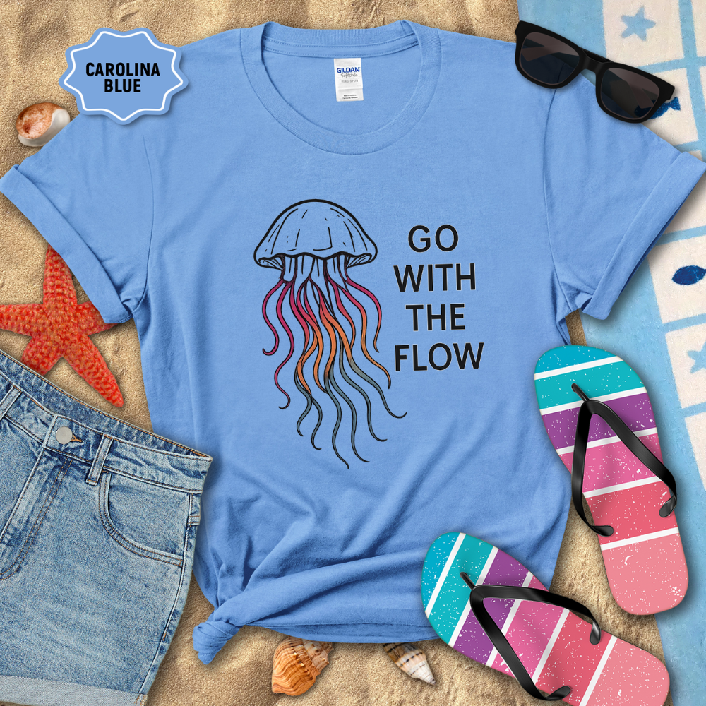 Blue t-shirt with jellyfish graphic and 'Go With The Flow' text, surrounded by beach items on sand.