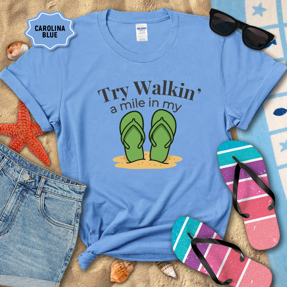 Blue t-shirt with flip-flop graphic and text, sunglasses, and beach accessories on sand.