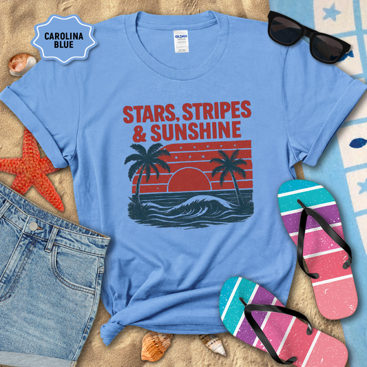 Blue t-shirt with 'Stars, Stripes & Sunshine' design on a sandy background with beach accessories.