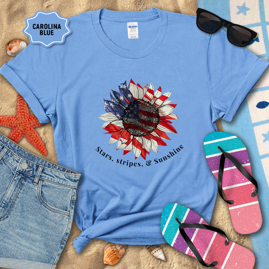 Blue t-shirt with a sunflower design and text, surrounded by beach accessories on sand.
