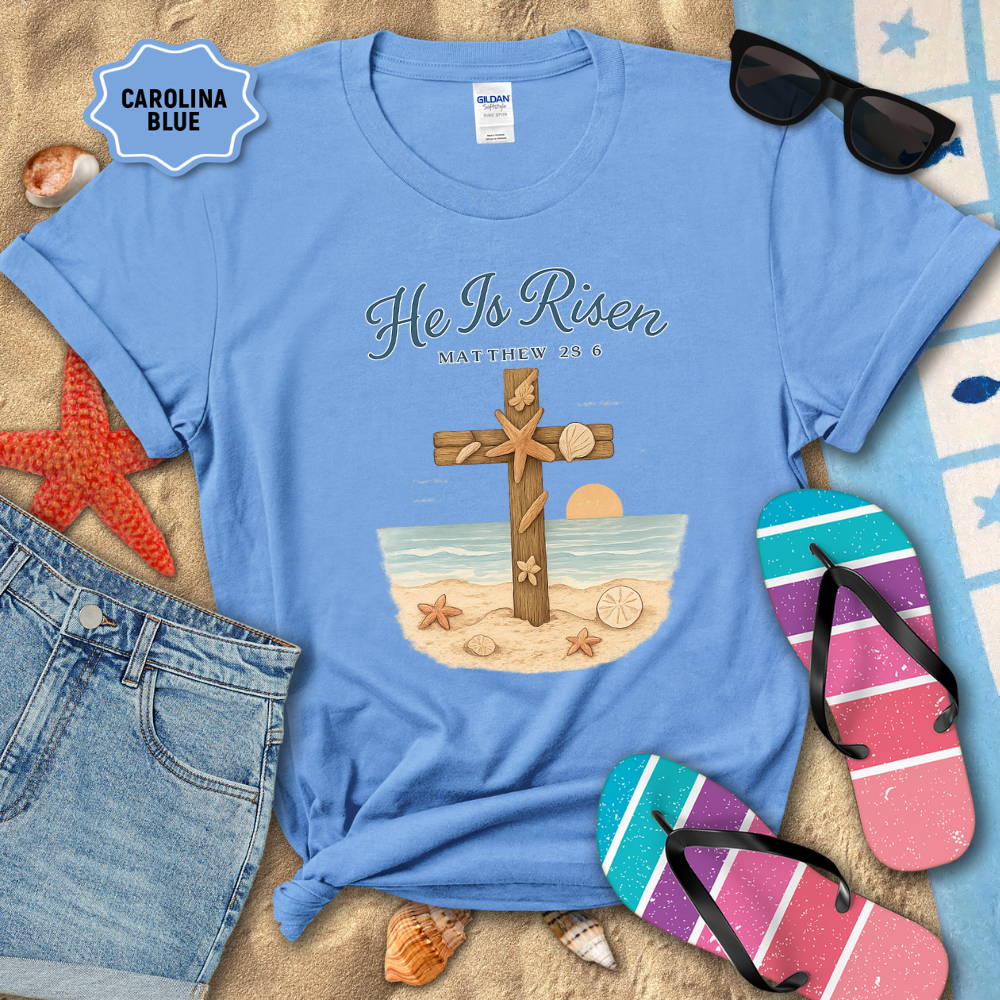 Blue t-shirt with 'He Is Risen' design on a sandy background with beach accessories.