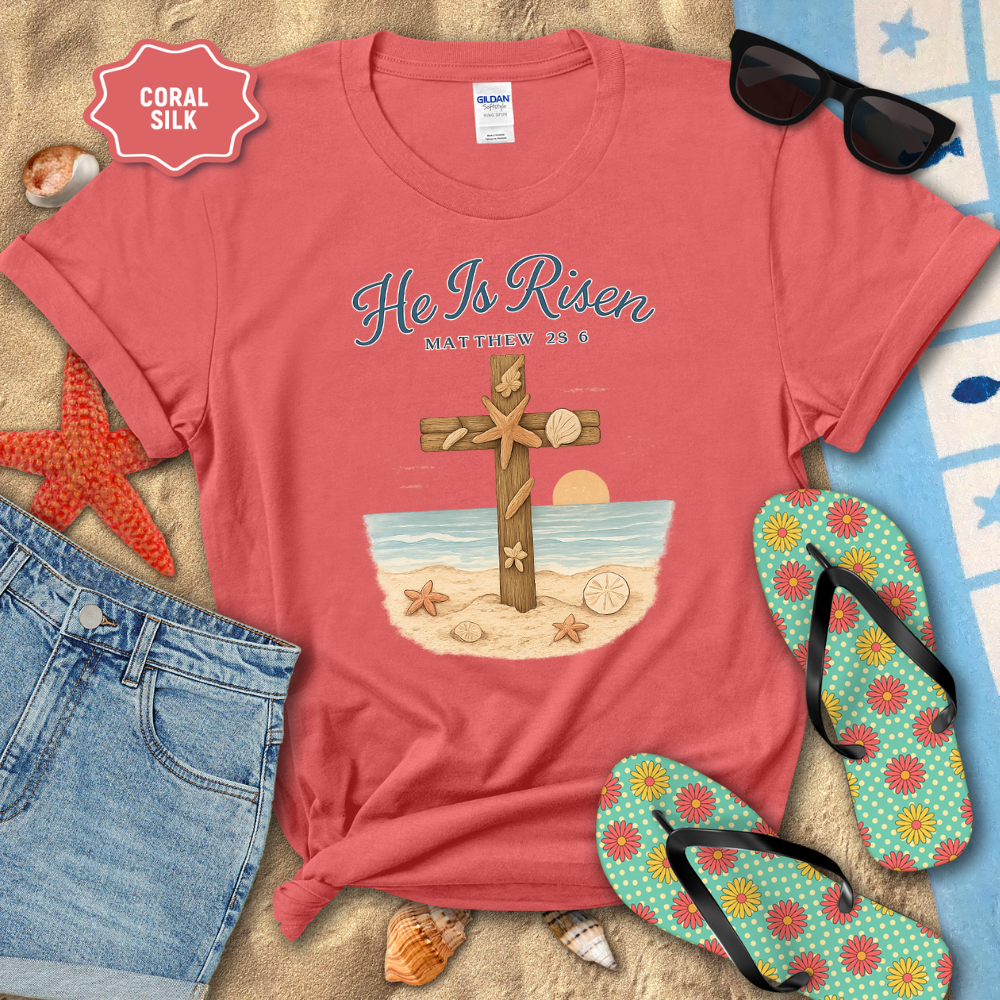 Coral t-shirt with 'He Is Risen' design on a sandy surface with beach accessories.