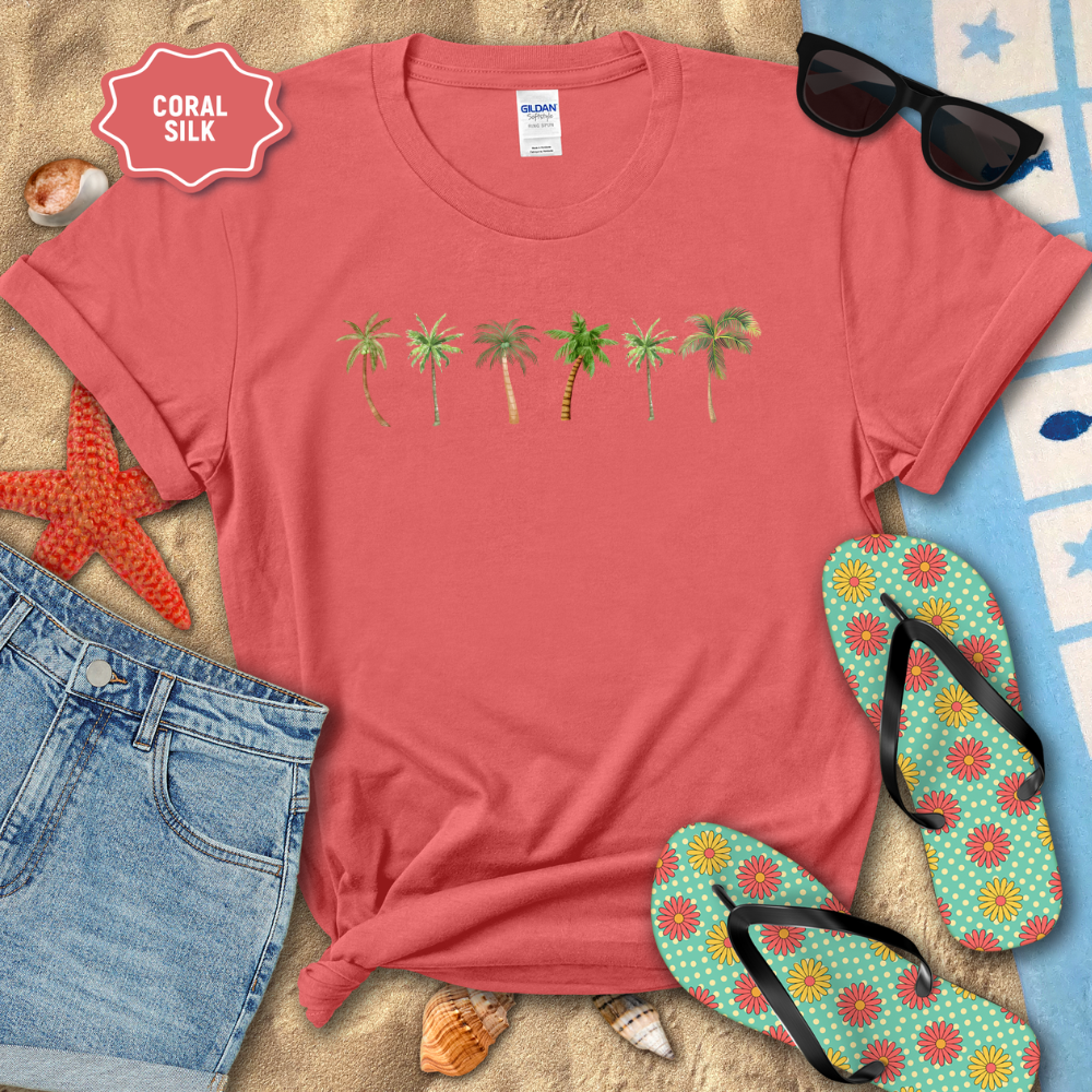 Coral t-shirt with palm tree design on a sandy surface with beach accessories.