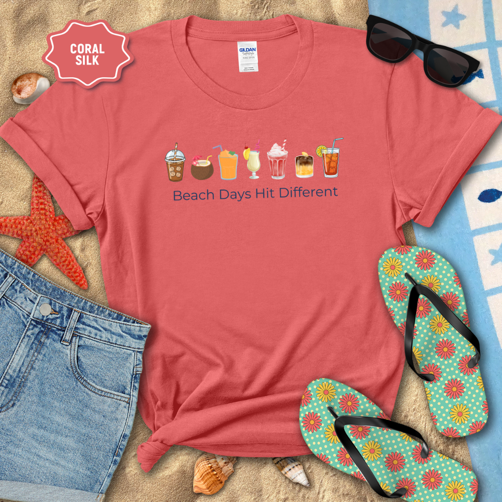 Coral t-shirt with beach-themed design on sand with sunglasses, flip-flops, and denim shorts.