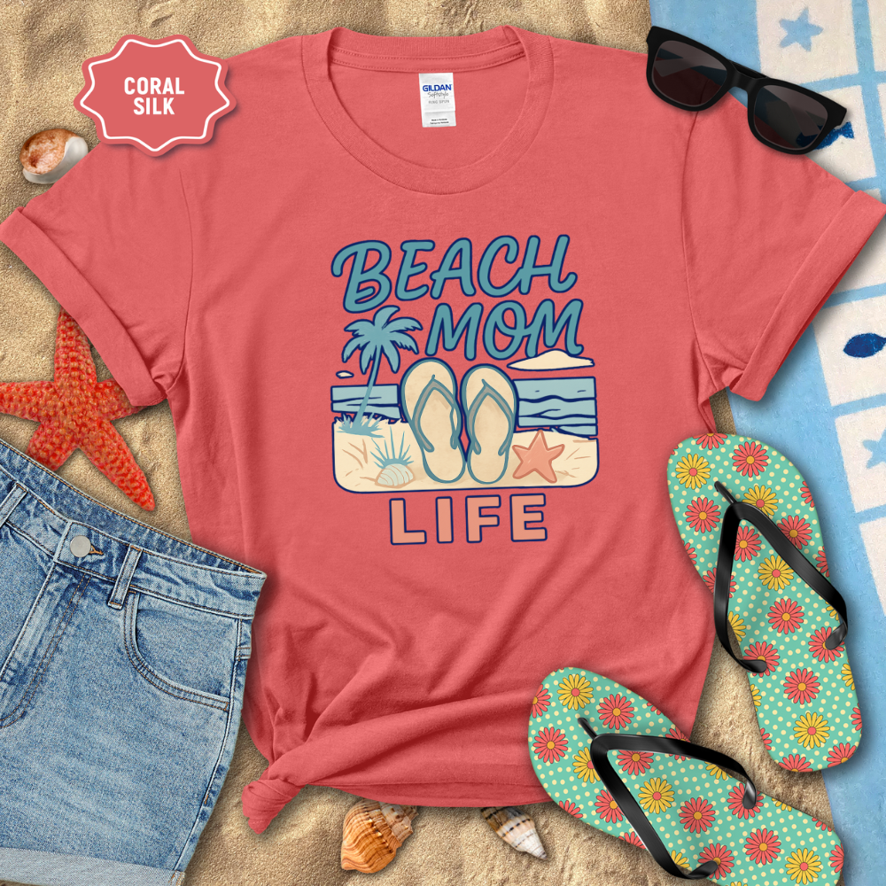 Coral t-shirt with 'Beach Mom Life' design on sand with sunglasses, flip-flops, and denim shorts.