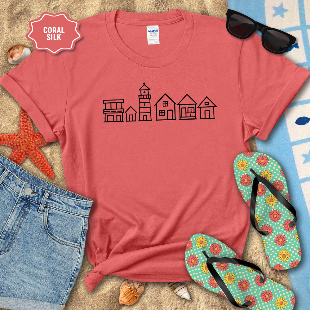 Red t-shirt with a house and lighthouse design, sunglasses, flip-flops, and beach items on sand.