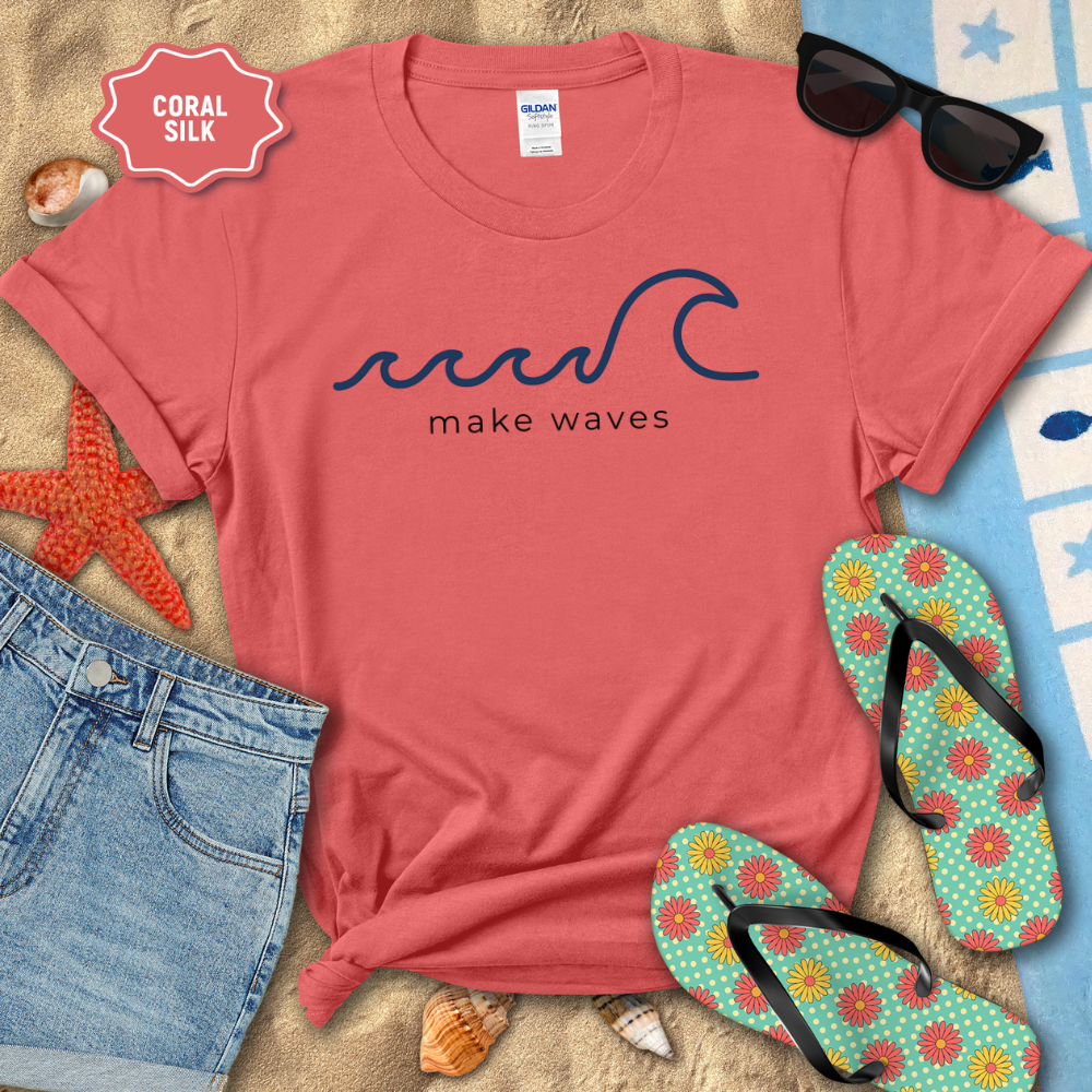 Coral t-shirt with 'make waves' design on a sandy surface with beach accessories.