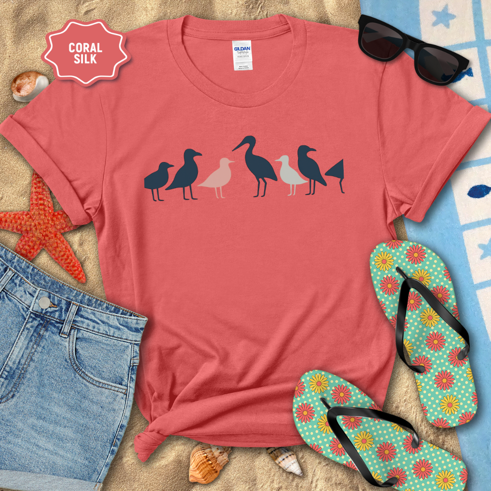 Coral t-shirt with bird design on a beach setting with sunglasses, flip-flops, and denim shorts.