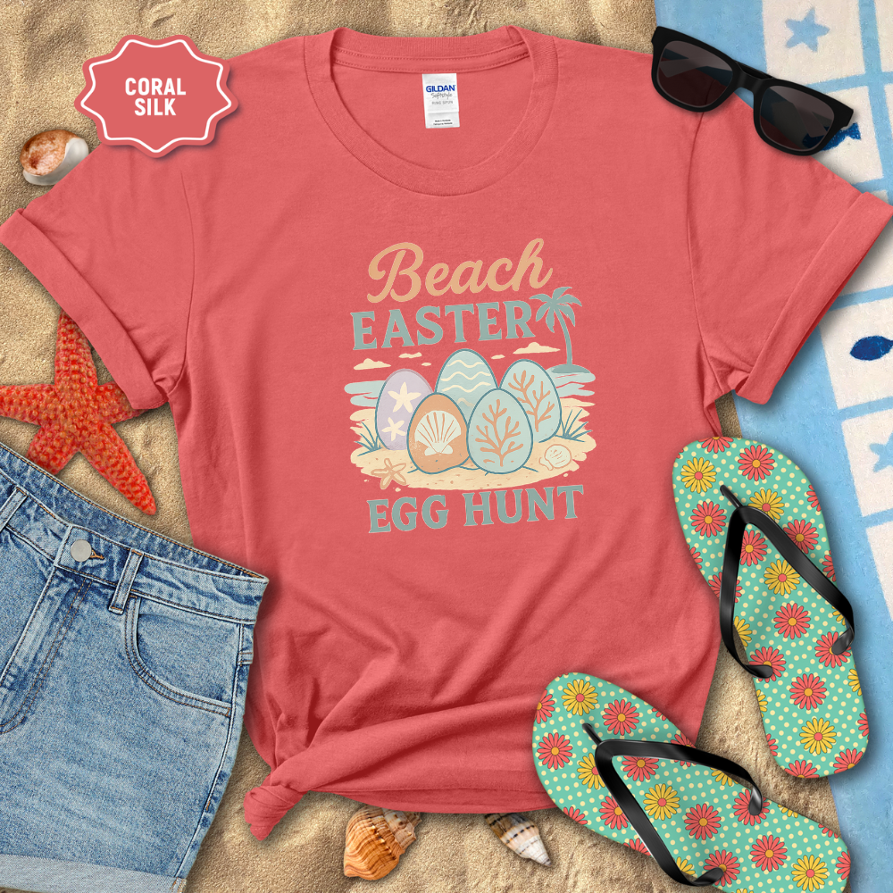 Coral t-shirt with 'Beach Easter Egg Hunt' design on a sandy surface with flip-flops and sunglasses.