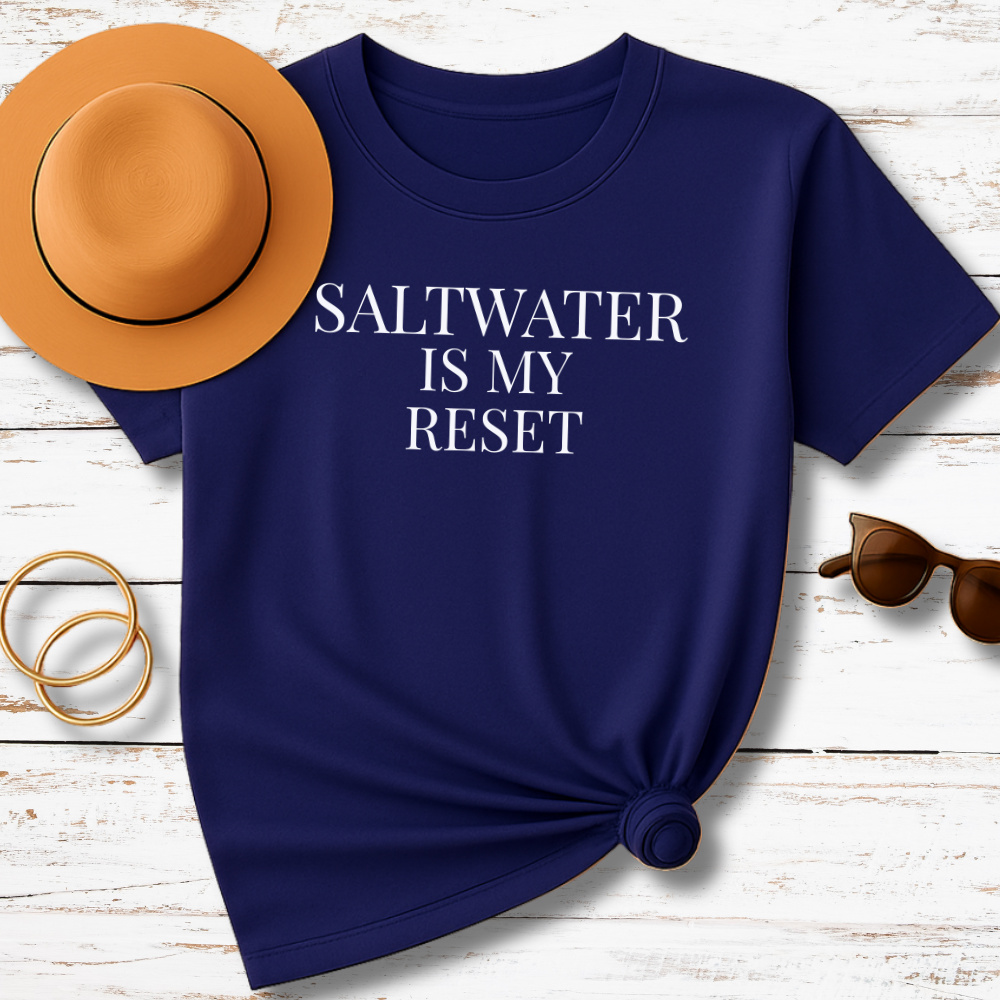 Navy blue t-shirt with 'SALTWATER IS MY RESET' text, accompanied by a hat, sunglasses, and gold rings on a wooden surface.