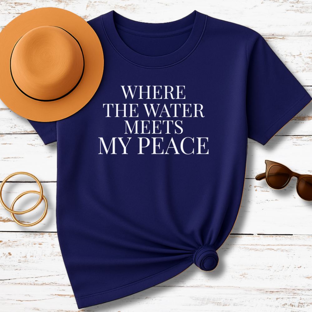 Navy blue t-shirt with 'Where the Water Meets My Peace' text, accompanied by a hat, sunglasses, and bracelets on a wooden surface.