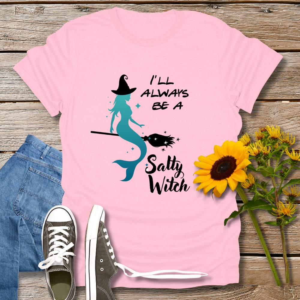 Pink t-shirt with mermaid and witch design, placed on a wooden surface with jeans, shoes, and sunflowers.