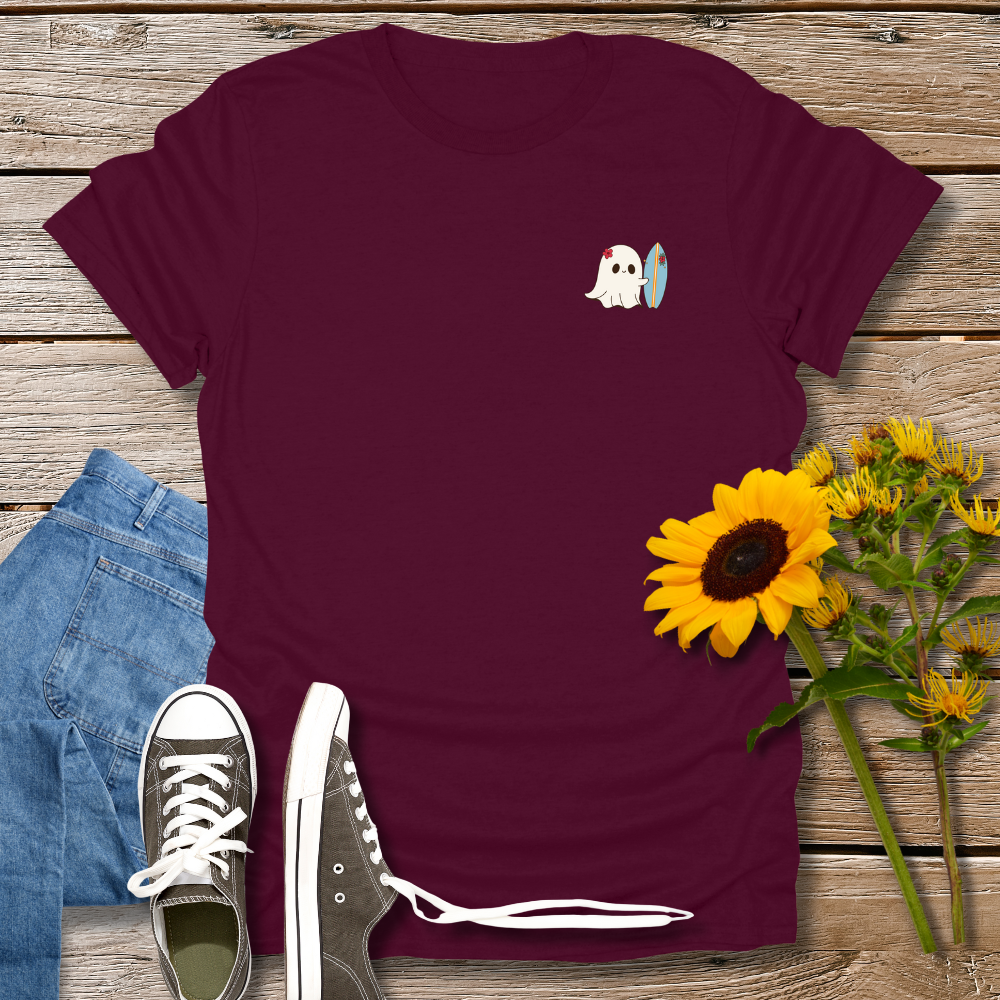 Maroon t-shirt with a ghost holding a surfboard design, paired with jeans and sneakers on a wooden surface.