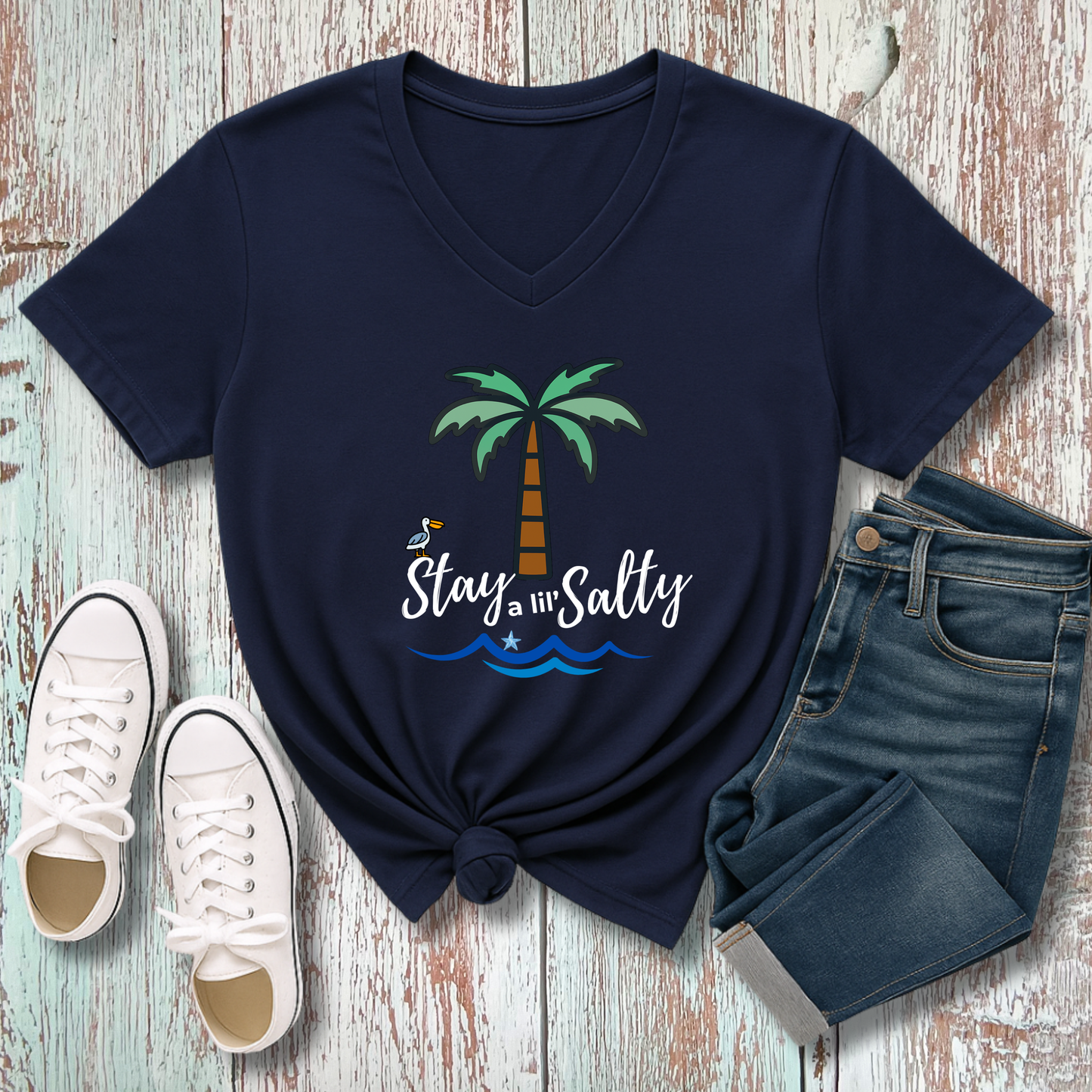 Navy blue t-shirt with palm tree and 'Stay Salty' text, paired with white sneakers and jeans on a wooden surface.