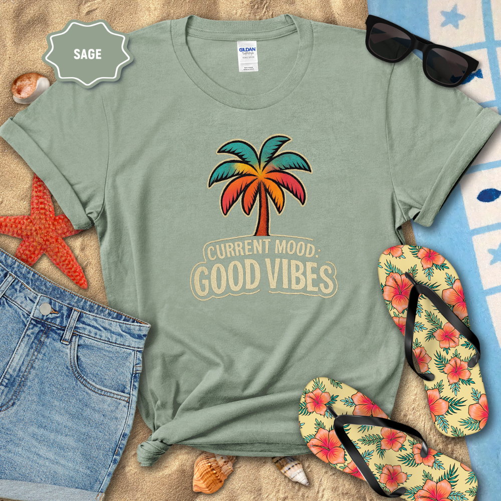 Sage green t-shirt with palm tree and 'Current Mood: Good Vibes' text, surrounded by beach accessories.