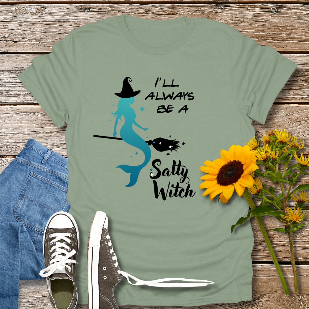 Green t-shirt with mermaid graphic and text 'I'll always be a salty witch' on a wooden surface with jeans, shoes, and sunflowers.